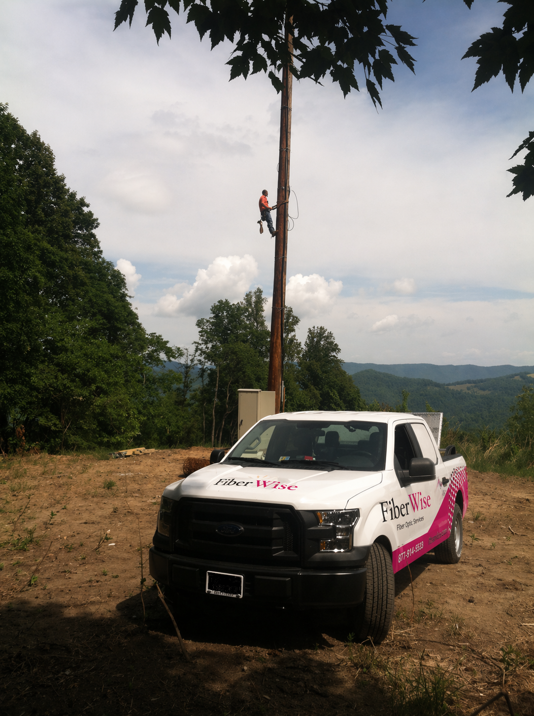 A FiberWise crew member climbing a pole on a ridgeline in Appalachia, FiberWise truck in the foreground